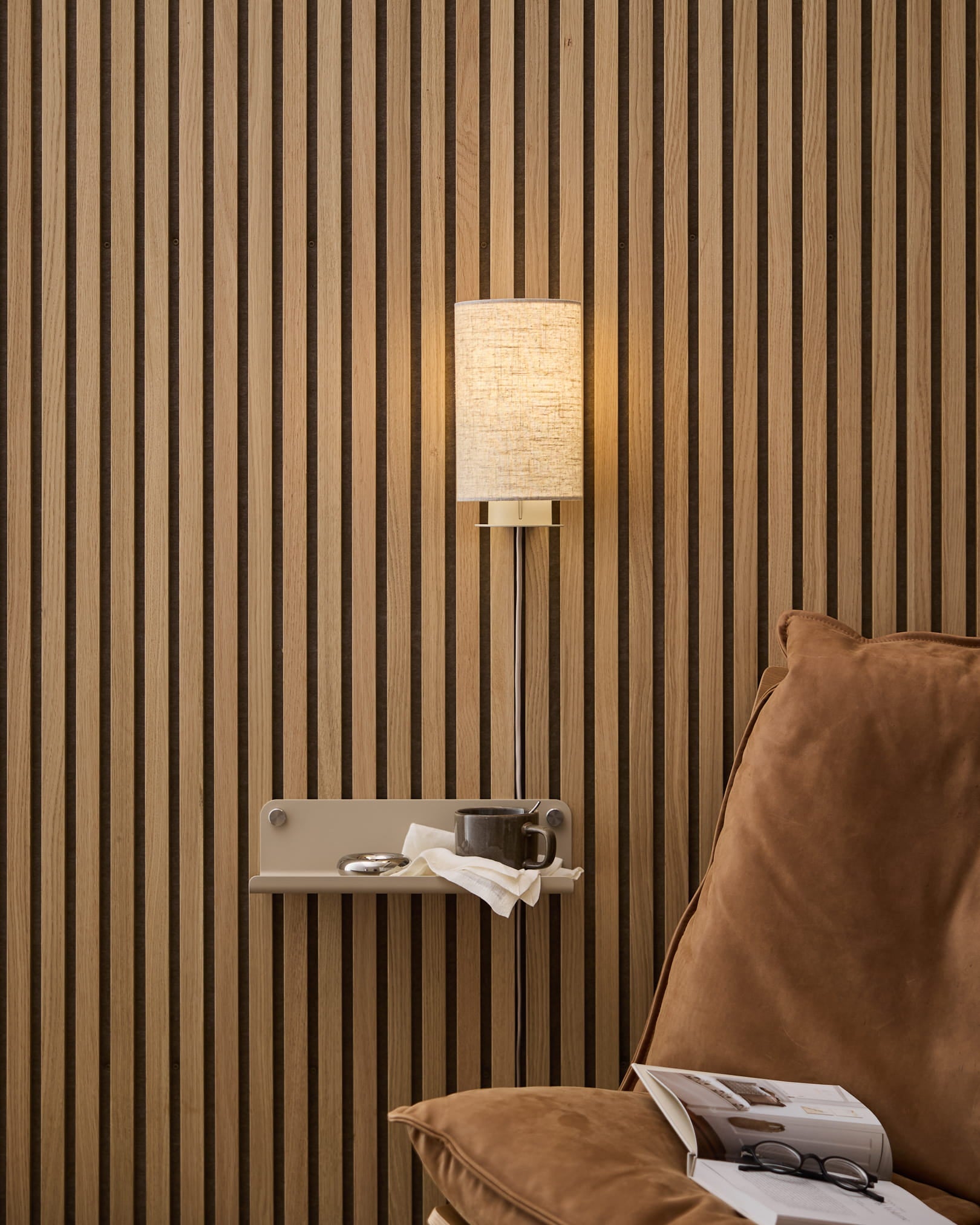 En beige WoodUpp Feya-væglampe i hør og bomuld monteret over en hyggelig lænestol og et sidebord med bog og kop mod et nordisk akustikpanel af trælameller.